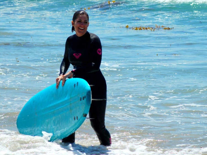 woman smiling in a wetsuit carrying a surfboard