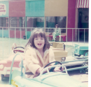 Young Karen Pery laughing as she is driving a toy car ride